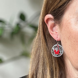 Close-up of a person wearing a silver earring with a red design, against a blurred natural background.
