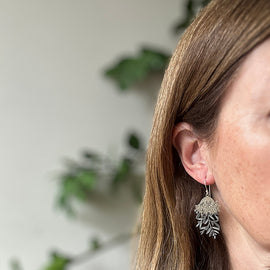 Close-up of a person wearing a silver leaf-shaped earring with a blurred green plant background.