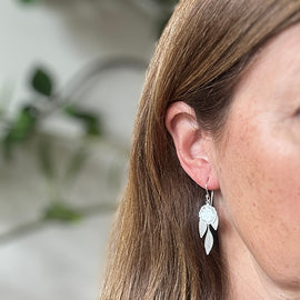 Close-up of a person wearing a silver earring with a leaf design against a blurred natural background.