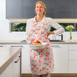 Woman in a kitchen wearing a floral apron, holding a plate of pancakes.